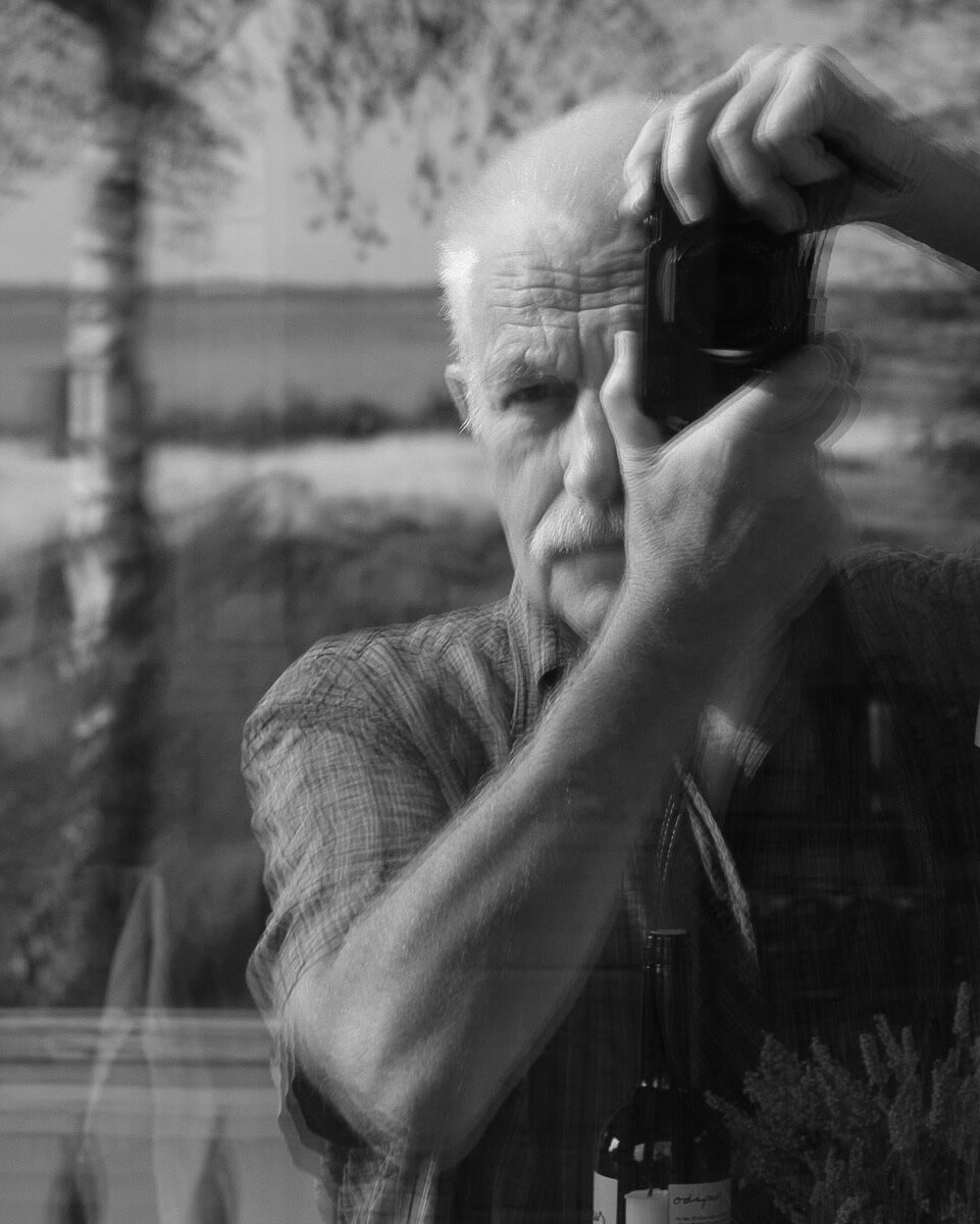 Schwarz-Weiß-Foto des Künstlers Uwe Greiß, der sich selbst porträtiert in einer spiegelnden Oberfläche und mit einem Strand und einem Birkenstamm im Hintergrund