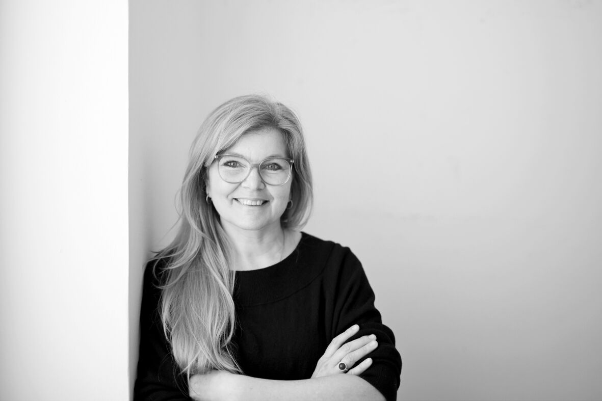 Schwarz-Weiß-Foto der Künstlerin Sonja Koczula, die mit verschränkten Armen, dunklem Oberteil und lächelnd an eine weiße Wand lehnt, sie trägt lange, helle Haare über die Schulter gelegt und eine Brille