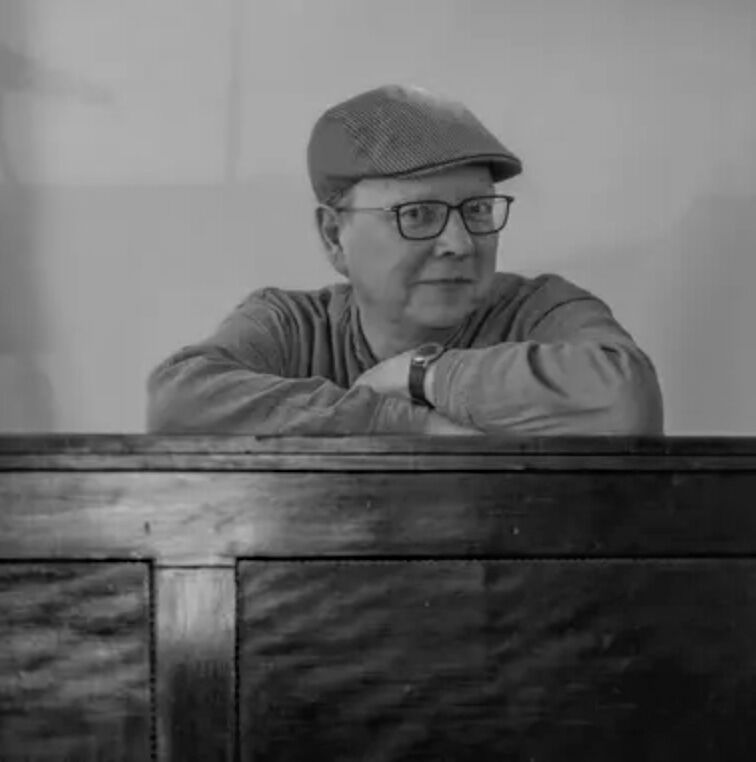 Schwarz-Weiß-Foto des Künstlers Martin Grieß, der mit den Armen auf einer Holzwand lehnt, Brille und Schiebermütze trägt