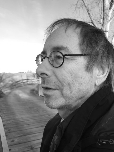Schwarz-Weiß-Foto des Künstlers Melvyn Poore im Profil vor einem Weg mit einer Birke, er trägt kurze Haare, eine Jacke und Brille
