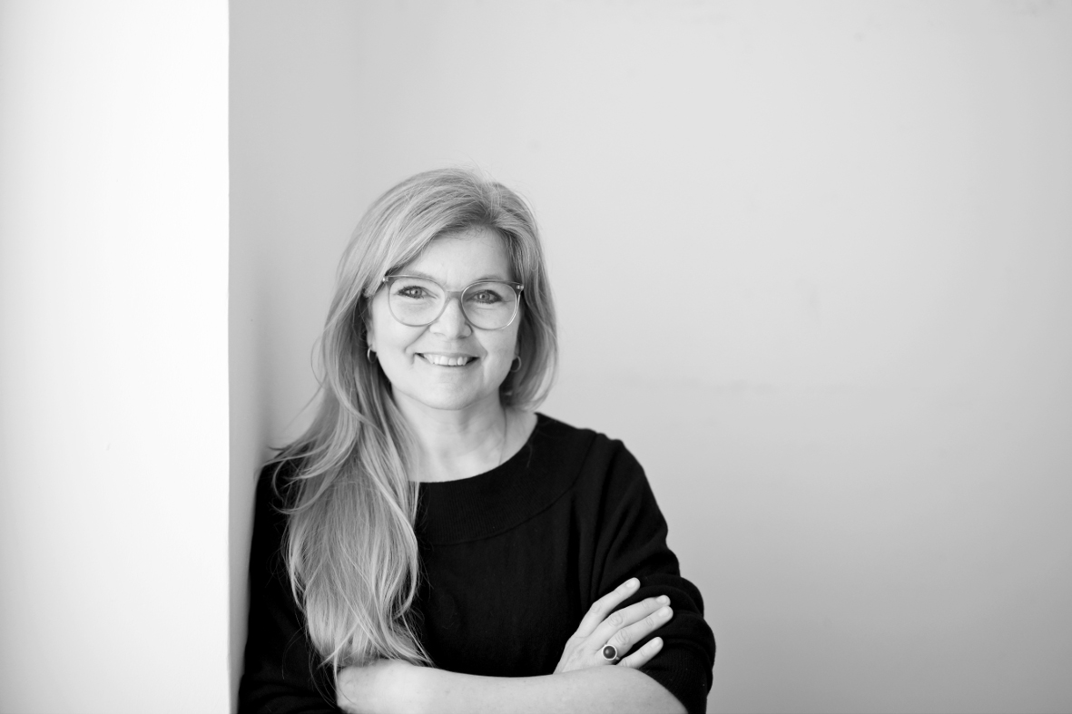Schwarz-Weiß-Foto der Künstlerin Sonja Koczula, die mit verschränkten Armen, dunklem Oberteil und lächelnd an eine weiße Wand lehnt, sie trägt lange, helle Haare über die Schulter gelegt und eine Brille
